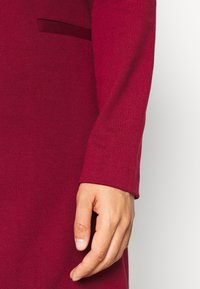 Robe bordeaux à manches longues avec une texture lisse, dotée d'une poche latérale et d'un design ajusté qui met en valeur la silhouette des bras.