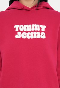 Un sweat à capuche fuchsia en tissu doux avec un logo "TOMMY JEANS" en relief blanc sur la poitrine. Inclut une capuche avec cordon de serrage.