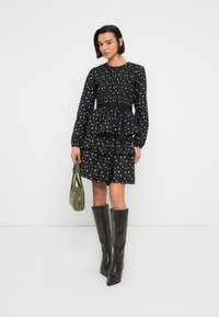 Robe fleurie noire avec des couches, des manches longues et une taille à nouer. Associée à des bottes marron montantes jusqu'au genou et à un sac à main vert.