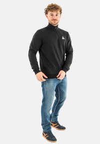 Zwarte zip-neck sweatshirt met ribbelmanchetten, gecombineerd met blauwe spijkerbroeken en donkere sneakers met bruine accenten.