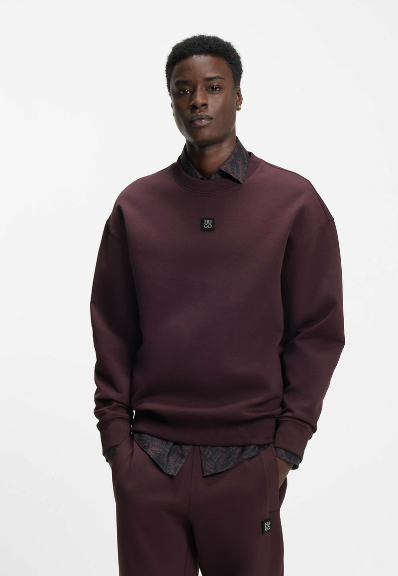 Burgundy sweatshirt with a round neck, ribbed cuffs, and a black logo patch; worn over a dark patterned shirt and matching pants.