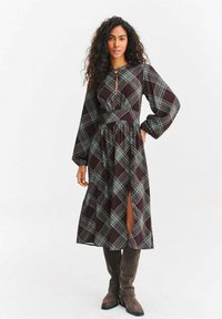 Długa sukienka w kratę w kolorach burgundu i zieleni, z dekoltem z wycięciem, długimi rękawami, paskiem w talii oraz rozcięciem po boku, wykonana z lekkiego materiału.