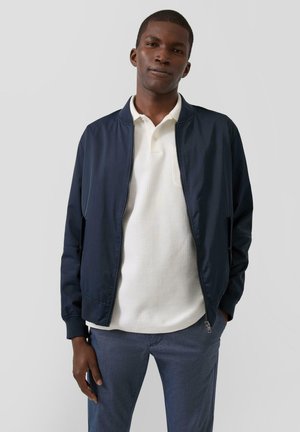 Giovane uomo con giacca bomber blu navy sopra una polo bianca e pantaloni blu, in piedi con una mano in tasca, su sfondo grigio.