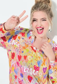Farverig gennemsigtig bluse med et abstrakt mønster med hjerter og ordet "FRED." Livlige nuancer af pink, rød, gul og blå.
