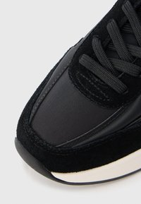 Musta tennari, joka on valmistettu mokasta ja synteettisistä materiaaleista, joissa on teksturoidut kengännauhat ja pehmustettu, pyöreä kärki. Minimalistinen muotoilu.