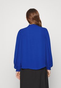 Blusa blu reale con maniche lunghe a sbuffo, texture liscia e colletto arricciato. L'orlo è leggermente arrotondato sul retro.