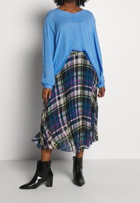 Lichtblauwe lange mouwen top met een v-hals, gecombineerd met een geplooide geruite rok in zwart, blauw, groen en wit. Zwarte enkellaarsjes met een spitse neus.