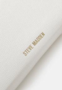 Gros plan sur du cuir blanc texturé avec le nom de la marque "STEVE MADDEN" en or embossé en diagonale.