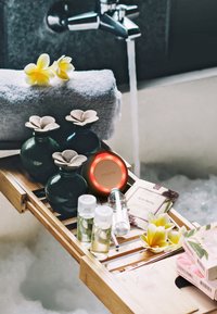 Bambusbadrack som håller tre gröna keramiska flaskor med blomlådor, en röd Foreo-enhet, olika små flaskor och förpackningar, med en handduk och blom-accenter.