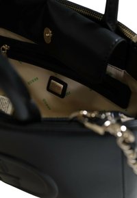 Borsa nera con esterno in pelle liscia. L'interno presenta una fodera beige con un motivo verde "GUESS", un compartimento con zip e una placca con il marchio.