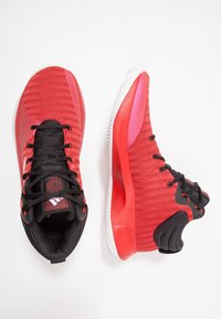 Chaussures de sport rouges avec une tige texturée, des lacets noirs et une semelle blanche. Elles disposent d'un col rembourré et d'un design flexible pour le confort.