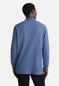 Hombre de espaldas vistiendo una camisa polo de manga larga azul y pantalones negros, frente a un fondo blanco liso.