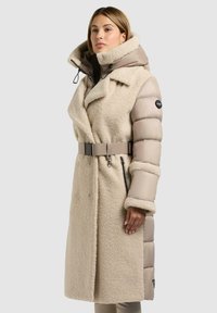 Beige jas van fluffy stof met een soepele puffer-mouw, een ceintuur in de taille, een grote capuchon en zijzakken met rits. Geaccentueerd met een logopatch.