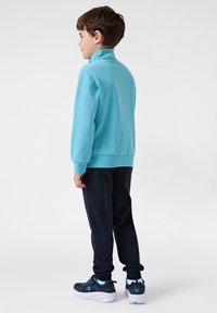 Felpa azzurra con colletto alto, abbinata a pantaloni blu scuro e scarpe sportive nere e blu, vista da dietro.