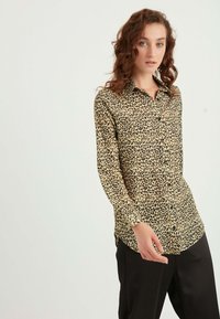 Leopardmønstret langærmet skjorte med knaplukning i beige og sort, med krave og lige snit, parret med sorte bukser.