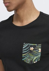 Camiseta negra de cuello redondo que presenta un bolsillo en el pecho izquierdo con un patrón de follaje verde y flores amarillas. Textura de algodón suave.