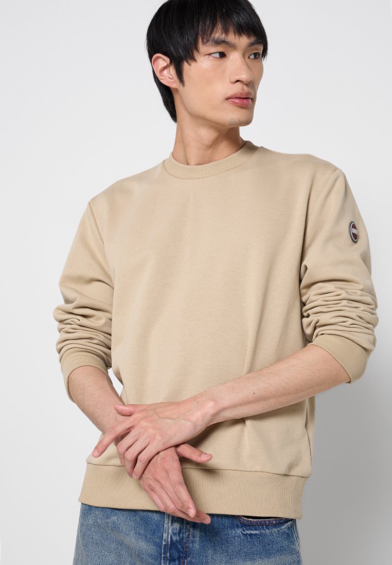 Felpa in cotone beige con collo rotondo, polsini e orlo a coste, dotata di una patch con il logo sulla manica sinistra. Texture liscia e vestibilità ampia.