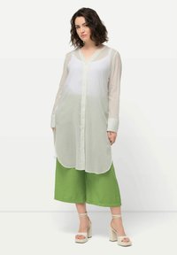 Leichte, durchscheinende weiße Bluse mit V-Ausschnitt, langen Ärmeln und Knopfverschluss. Kombiniert mit grünen weit geschnittenen Hosen und klobigen Sandalen mit Absatz.