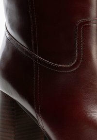 Burgundowy skórzany botek za kostkę o gładkiej fakturze, z wysokim obcasem typu słupek i ozdobnym przeszyciem wzdłuż szwu i krawędzi.