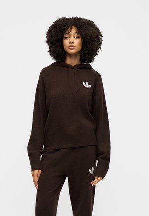 Donna con capelli ricci che indossa felpa con cappuccio e pantaloni marrone scuro abbinati con loghi bianchi Adidas, in piedi davanti a uno sfondo chiaro e semplice.