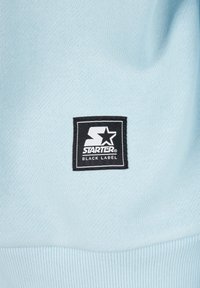 Light blue sweatshirt featuring a black rectangular tag with white "STARTER" text and a star logo, set against a textured fabric.