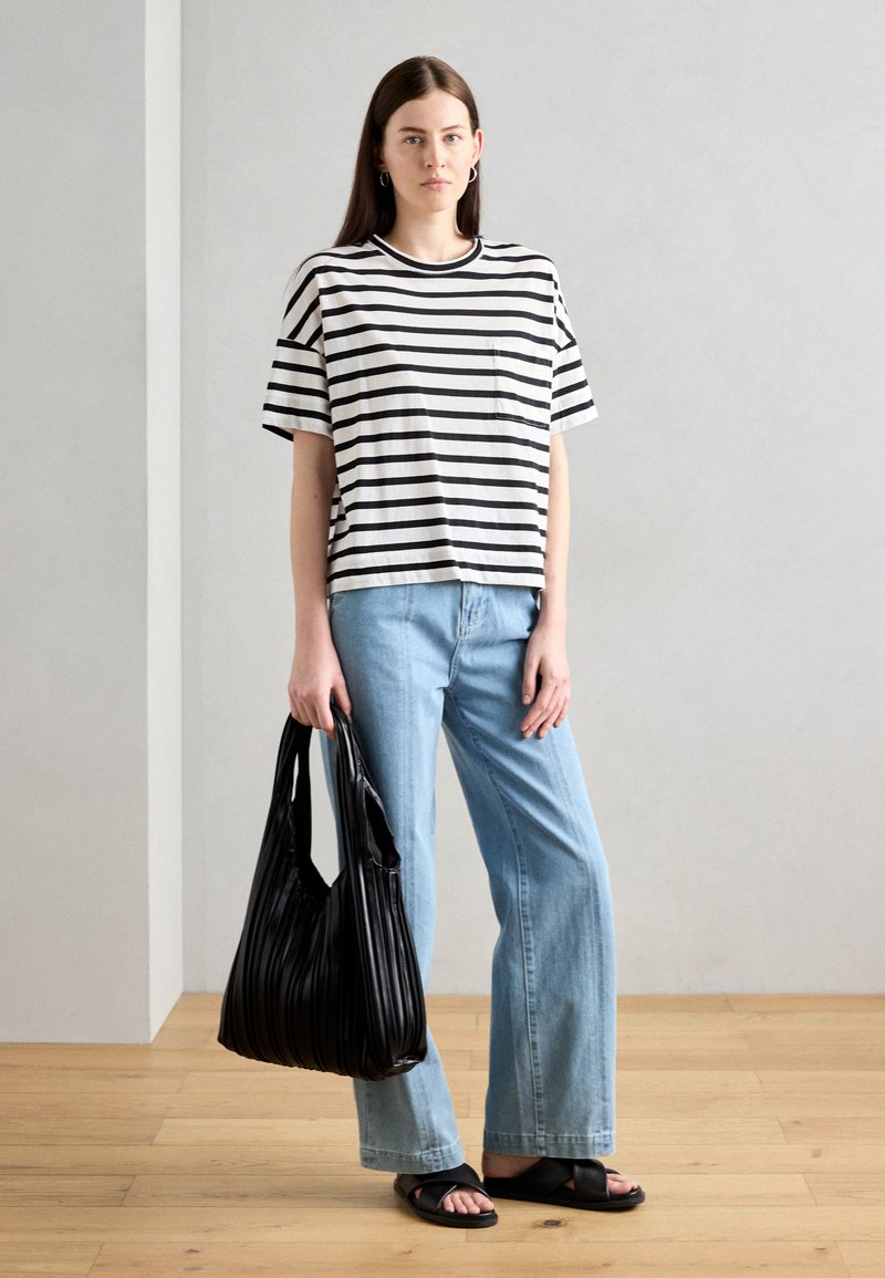 Gestreiftes Schwarz-Weiß-T-Shirt, hellblaue weit geschnittene Jeans, schwarze plissierte Tragetasche und schwarze Sandalen. Das Model steht vor einem grauen Hintergrund.