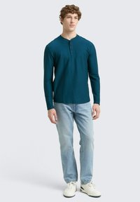 Türkise Langarm-Henley-Shirt mit drei Knöpfen, kombiniert mit hellblauen Jeans und weißen Sneakers auf einem neutralen Hintergrund.