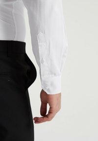 Chemise blanche à manches longues avec une texture lisse, dotée d'un poignet pointu avec fermeture à bouton, portée avec un pantalon noir.