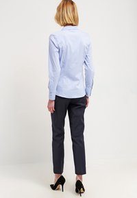 Camicia azzurra chiara con colletto realizzata in tessuto liscio, abbinata a pantaloni sartoriali blu scuro. Tacchi alti neri completano l'outfit visto da dietro.