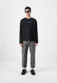 Camicia nera a maniche lunghe con logo bianco, abbinata a pantaloni grigi con motivo floreale. Gli accessori includono occhiali da sole scuri e scarpe nere.