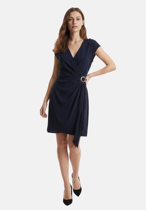 Donna in piedi, che indossa un vestito a portafoglio a maniche corte blu navy con un dettaglio ad anello d'argento in vita e décolleté nere con punta appuntita.