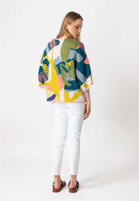 Multicolored gebreide top met brede mouwen, voorzien van abstracte vormen in groen, blauw, roze en geel. Gemaakt met witte broek en sandaaltjes.