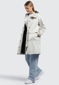 Impermeabile leggero in crema con cappuccio, tasche frontali e bottoni a pressione, abbinato a jeans azzurri e sneakers bianche.