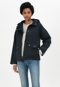 Veste imperméable marine avec capuche, dotée de quincaillerie dorée, de deux poches avant et d'un cordon de serrage au niveau du cou. Modèle portant un pull léger et un jean.