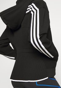 Sudadera negra con capucha que presenta textura canalé, rayas blancas en las mangas y un dobladillo tipo peplum. La tela tiene un acabado suave y un diseño estructurado.