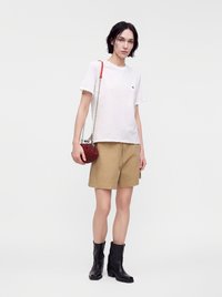 Γυναίκα με λευκό T-shirt, ταμπά σορτς, μαύρα μποτάκια μέχρι τον αστράγαλο, κρατώντας μια μικρή κόκκινη τσάντα με λουράκι αλυσίδα σε ουδέτερο φόντο.