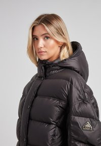 Giovane donna con capelli biondi che indossa un giubbotto imbottito con cappuccio nero, con un patch con logo sulla manica, che guarda leggermente di lato.