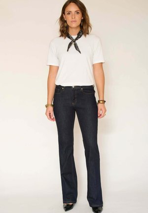 Jean évasé bleu foncé en denim, associé à un t-shirt blanc uni et une écharpe noire. Comprend des poches et des détails de couture.