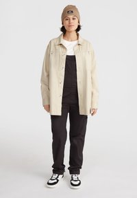 Chaqueta de lona beige sobre una camisa blanca, petos negros y zapatillas de deporte en blanco y negro. Incluye un gorro de punto color beige con un parche de logo.