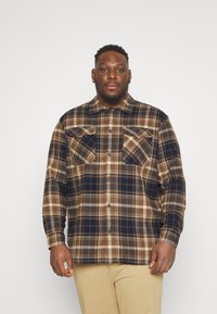 Camisa de franela de cuadros marrón y marino con dos bolsillos en el pecho, botones y un cuello con solapa. La tela parece suave y texturizada.