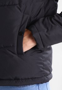 Giacca a vento nera con una tasca con zip e chiusura a scatto sul lato, caratterizzata da una texture trapuntata e maniche aderenti.