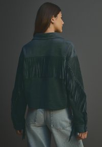 Kurzjacke aus grünen Wildleder mit Fransenverzierungen auf dem Rücken und den Ärmeln, ausgestattet mit einem Reverskragen und knöpfbaren Manschetten. Getragen mit hellen Jeans.