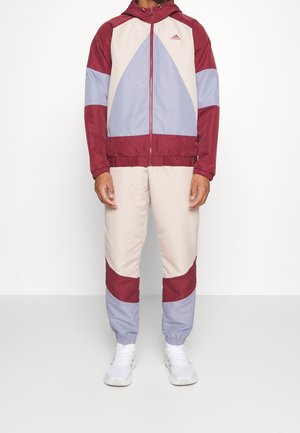 Mann trägt einen Adidas-Trainingsanzug in Bordeaux, Helllila und Beige mit weißen Turnschuhen, steht vor einer schlichten weißen Wand.