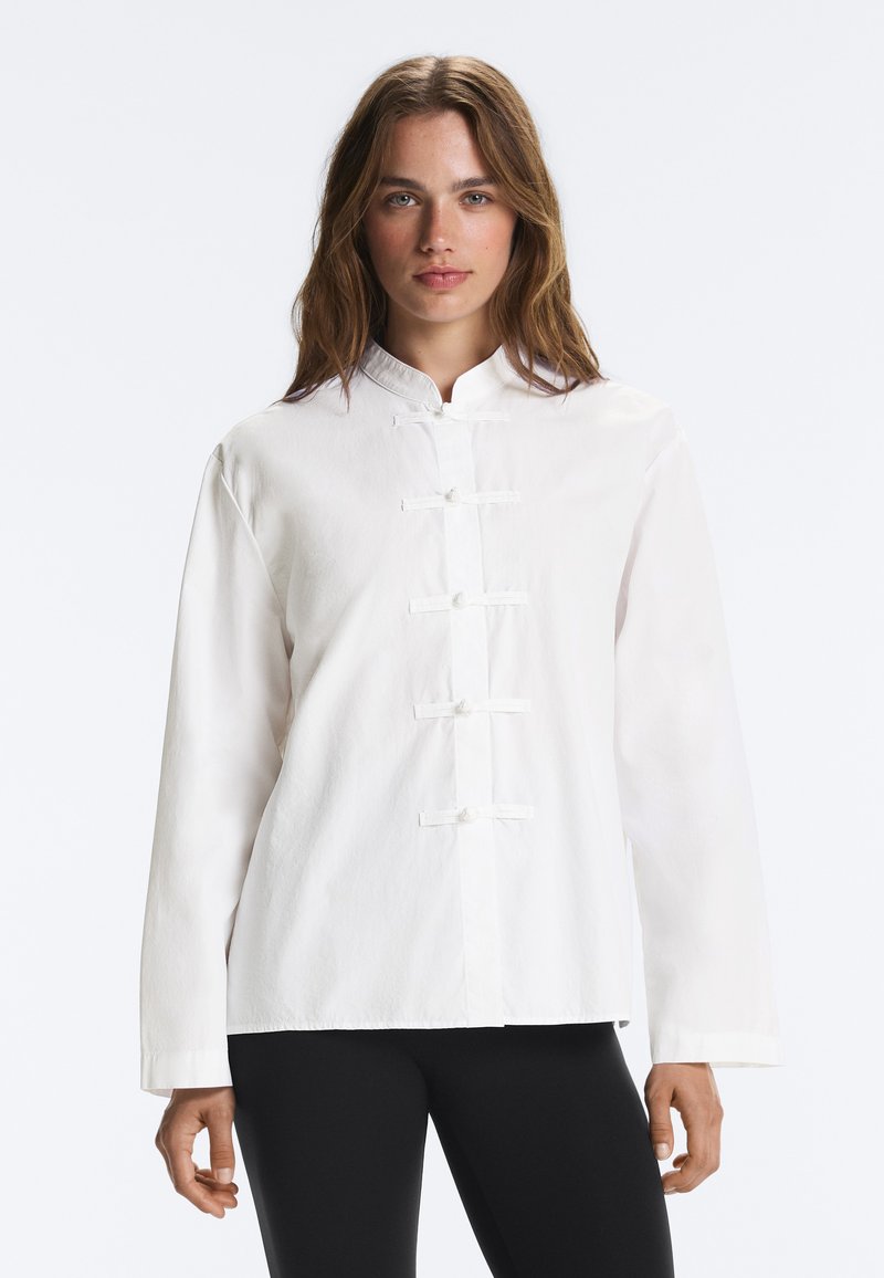 Jeune femme aux cheveux bruns ondulés portant une chemise blanche boutonnée de style traditionnel et un pantalon noir, debout devant un fond uni.