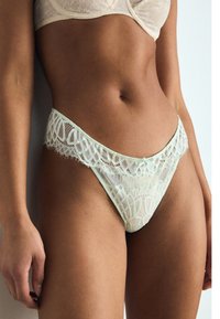 Lace bikini in lichtgroen met gegolfde randen, voorzien van intricate patronen en een klein strikje aan de voorkant, gemaakt van doorschijnende stof.