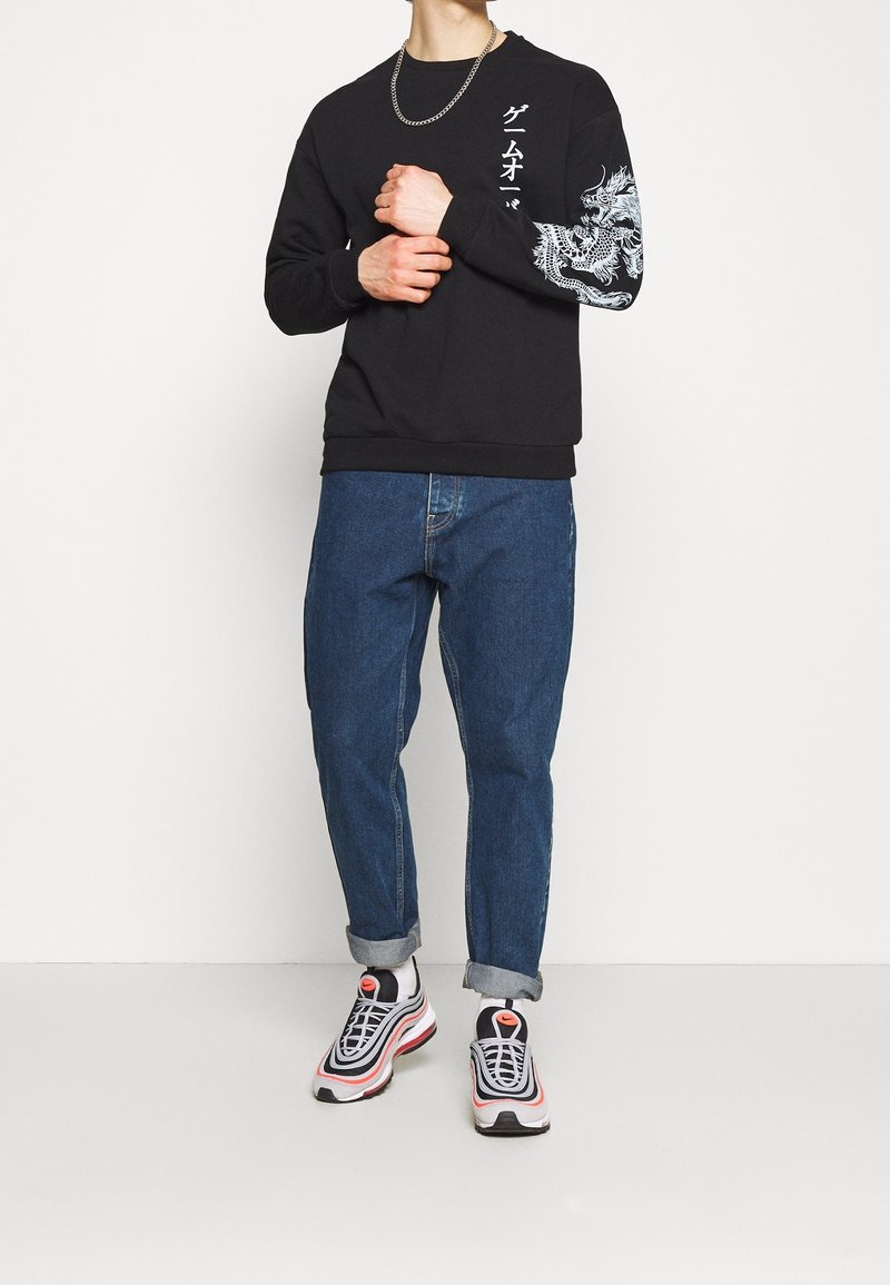 Homme portant un sweat-shirt noir avec un motif de dragon blanc sur la manche et du texte japonais, jean bleu retroussé, baskets blanches avec des accents rouges.