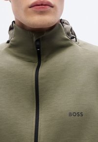Μπουφάν με κουκούλα σε ελιά πράσινο, με μπροστινό μαύρο φερμουάρ, μαλακή υφή και μικρό λογότυπο "BOSS" στην αριστερή πλευρά.