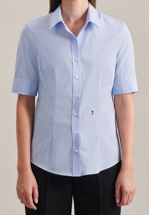 Chemise bleu clair à manches courtes, boutonnée avec col et petite rose noire brodée près du bouton inférieur, portée avec un pantalon noir.