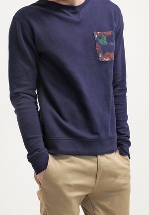 Mann trägt dunkelblauen Pullover mit Blumendruck auf der Brusttasche und beige Hose, Hände in den Taschen, vor schlichtem Hintergrund.