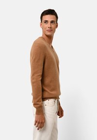 Hellbrauner, gestrickter Pullover mit V-Ausschnitt und langen Ärmeln. Strukturiertes Material, enganliegendes Design, kombiniert mit hellen Hosen.
